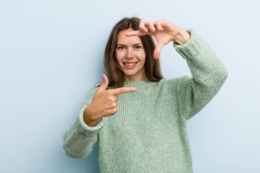 Genç ve güzel bir kadın mutlu, arkadaş canlısı ve pozitif hissediyor, gülümsüyor ve elleriyle bir portre veya fotoğraf çerçevesi yapıyor.