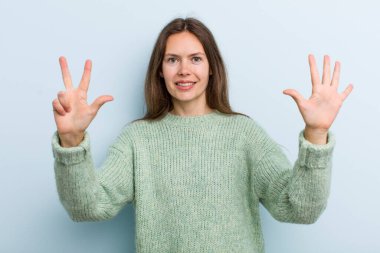 Genç ve güzel bir kadın gülümsüyor ve arkadaş canlısı görünüyor, 8 ya da 8 numarayı gösteriyor, elleri önde, geriye doğru sayıyor.