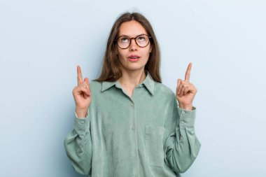 young adult pretty woman looking shocked, amazed and open mouthed, pointing upwards with both hands to copy space
