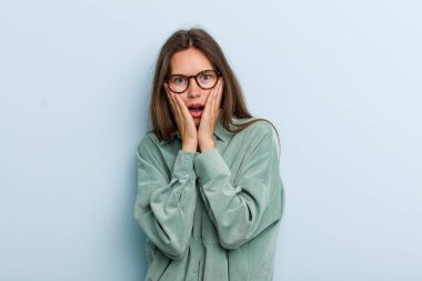 Genç ve güzel bir kadın şok olmuş ve korkmuş hissediyor, ağzı açık ve elleri yanaklarında korkmuş görünüyor.