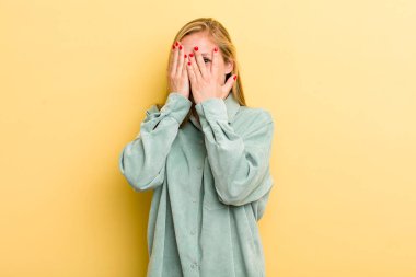 Genç, sarışın, güzel bir kadın. Elleriyle yüzünü kapatıyor. Şaşırmış bir ifadeyle parmaklarının arasından bakıyor.