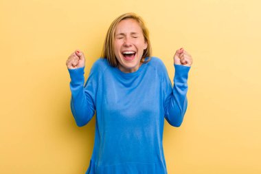 young adult blonde pretty woman looking extremely happy and surprised, celebrating success, shouting and jumping