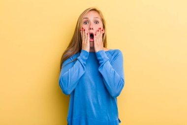 young adult blonde pretty woman feeling shocked and scared, looking terrified with open mouth and hands on cheeks