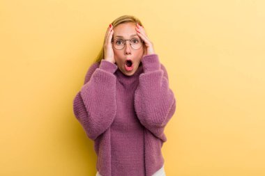 young adult blonde pretty woman feeling horrified and shocked, raising hands to head and panicking at a mistake