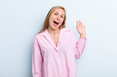 young adult blonde pretty woman smiling happily and cheerfully, waving hand, welcoming and greeting you, or saying goodbye