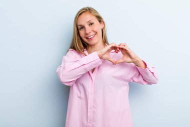 Genç, sarışın, güzel bir kadın gülümsüyor ve mutlu, sevimli, romantik ve aşık, iki eliyle kalbi şekillendiriyor.