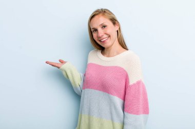 young adult blonde pretty woman smiling cheerfully, feeling happy and showing a concept in copy space with palm of hand