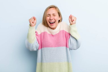 young adult blonde pretty woman looking extremely happy and surprised, celebrating success, shouting and jumping
