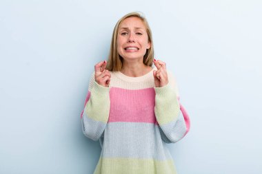 young adult blonde pretty woman crossing fingers anxiously and hoping for good luck with a worried look