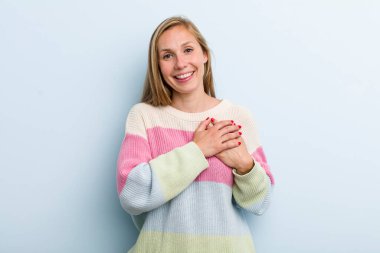 young adult blonde pretty woman feeling romantic, happy and in love, smiling cheerfully and holding hands close to heart
