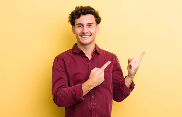 young handsome man smiling happily and pointing to side and upwards with both hands showing object in copy space