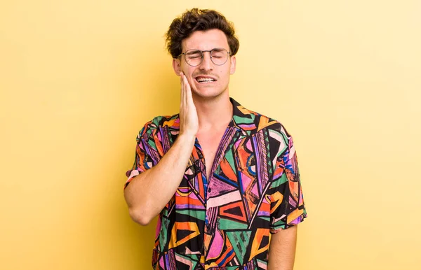 Genç yakışıklı adam yanağını tutuyor ve ağrılı diş ağrısı çekiyor, hasta, sefil ve mutsuz hissediyor, dişçi arıyor.