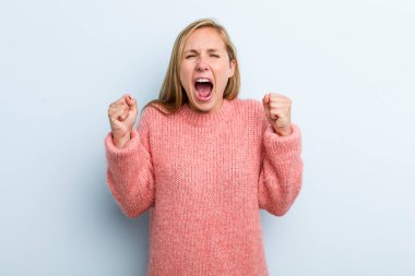young adult blonde pretty woman shouting aggressively with an angry expression or with fists clenched celebrating success