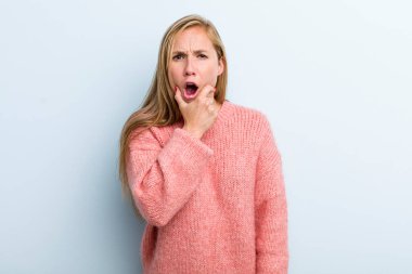 young adult blonde pretty woman with mouth and eyes wide open and hand on chin, feeling unpleasantly shocked, saying what or wow