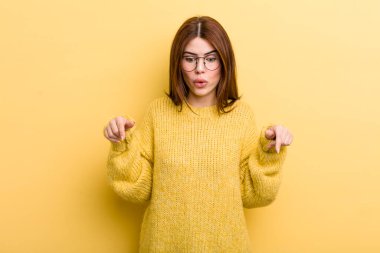 young adult pretty woman feeling shocked, open-mouthed and amazed, looking and pointing downwards in disbelief and surprise