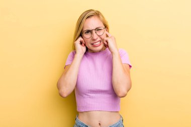 Genç, sarışın bir kadın sinirli, stresli ve sinirli görünüyor, iki kulağını da sağır edici bir ses, ses ya da yüksek sesle kapatıyor.