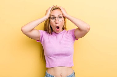 Genç sarışın kadın heyecanlı ve şaşırmış görünüyor, ağzı açık, iki eli başında, kendini şanslı hissediyor.