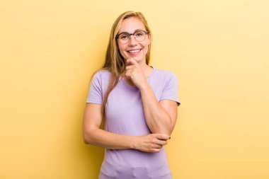 Genç, sarışın kadın mutlu görünüyor ve gülümsüyor. Elleri çenesinde, merak ediyor ya da soru soruyor. Seçenekleri karşılaştırıyor.