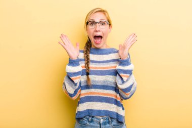 young adult  blonde woman looking happy and excited, shocked with an unexpected surprise with both hands open next to face