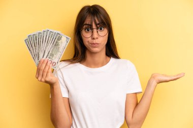 hispanic woman feeling puzzled and confused, scratching head. dollar banknotes concept