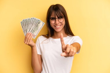 hispanic woman smiling proudly and confidently making number one. dollar banknotes concept