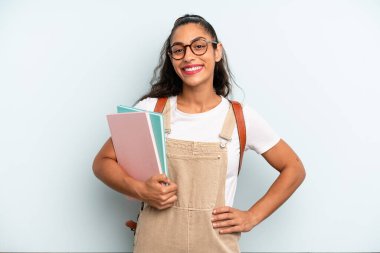 hispanic woman smiling happily with a hand on hip and confident. university student concept