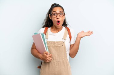 hispanic woman looking surprised and shocked, with jaw dropped holding an object. university student concept