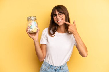 hispanic woman smiling confidently pointing to own broad smile. candies bottle concept