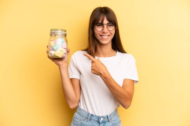 hispanic woman smiling cheerfully, feeling happy and pointing to the side. candies bottle concept