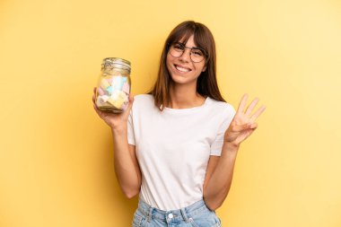 hispanic woman smiling and looking friendly, showing number three. candies bottle concept