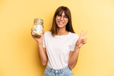 hispanic woman smiling and looking friendly, showing number two. candies bottle concept