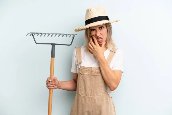 blonde woman with mouth and eyes wide open and hand on chin. farmer and rake cocnept