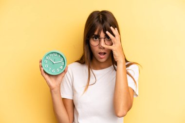 hispanic woman looking shocked, scared or terrified, covering face with hand. alarm clock concept