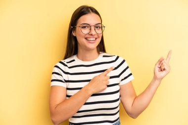 young adult pretty woman smiling happily and pointing to side and upwards with both hands showing object in copy space