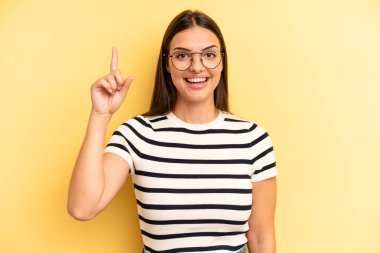 young adult pretty woman smiling cheerfully and happily, pointing upwards with one hand to copy space