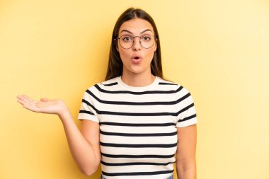 young adult pretty woman looking surprised and shocked, with jaw dropped holding an object with an open hand on the side