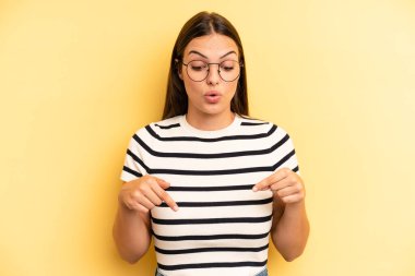 young adult pretty woman with open mouth pointing downwards with both hands, looking shocked, amazed and surprised