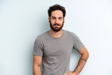 Genç yetişkin sakallı adam mutlu bir şekilde gülümsüyor, eli kalçasında ve kendinden emin, olumlu, gururlu ve arkadaş canlısı.