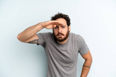 Genç, sakallı bir adam şaşkınlık ve şaşkınlık içinde bakıyor. Alnı açık, uzaklara bakıyor, izliyor ya da arıyor.