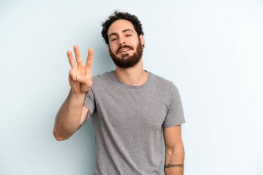 Genç, sakallı bir adam gülümsüyor ve arkadaş canlısı görünüyor. Elleri önde ve geriye doğru sayıyor.