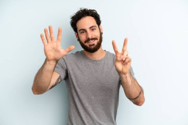 Genç, sakallı bir adam gülümsüyor ve dost canlısı görünüyor, 7 ya da 7 numarayı gösteriyor, eli önde, geriye doğru sayıyor.