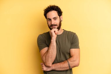 Genç, sakallı bir adam mutlu görünüyor ve eli çenesinde gülümsüyor, merak ediyor ya da soru soruyor, seçenekleri karşılaştırıyor.