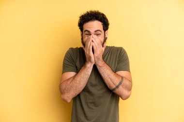 Genç sakallı adam mutlu, neşeli, şanslı ve şaşkın görünüyor. İki eliyle ağzını kapatıyor.