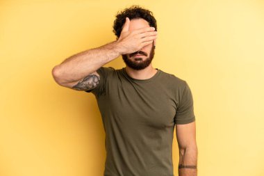 Genç, sakallı bir adam tek eliyle gözlerini kapatıyor korkmuş ya da endişeli hissediyor, merak ediyor ya da körü körüne bir sürpriz bekliyor.