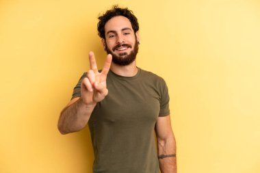 Genç, sakallı adam gülümsüyor ve arkadaş canlısı görünüyor. İki ya da iki numarayı gösteriyor. Elleri önde, geriye doğru sayıyor.