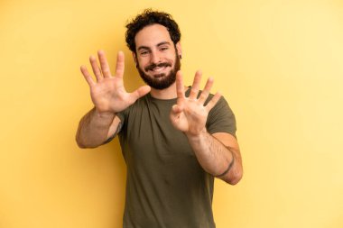 Genç, sakallı bir adam gülümsüyor ve dost canlısı görünüyor. 9 ya da 9 numarayı gösteriyor. Eli önde, geriye doğru sayıyor.