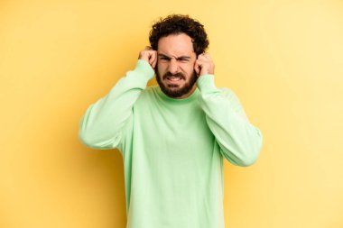 Genç, sakallı bir adam sinirli, stresli ve sinirli görünüyor, iki kulağını da sağır edici bir ses, ses ya da yüksek sesle kapatıyor.
