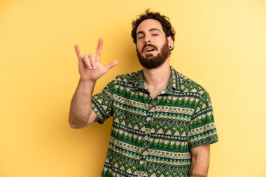 Genç sakallı adam mutlu, eğlenceli, kendinden emin, pozitif ve asi hissediyor, elle kaya ya da heavy metal işareti yapıyor.