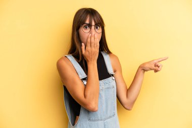 İspanyol güzel kadın kendini mutlu, şaşkın ve şaşırmış hissediyor. Ağzını eliyle kapatıyor ve yanal kopya alanını işaret ediyor.