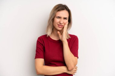 young adult blonde woman holding cheek and suffering painful toothache, feeling ill, miserable and unhappy, looking for a dentist